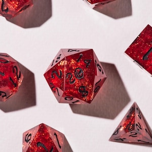 May include: A selection of red, multi-sided dice with black numbering. The dice have a glittery, speckled appearance. The dice are arranged on a white surface, with shadows cast from an overhead light source. The dice are used for tabletop games.