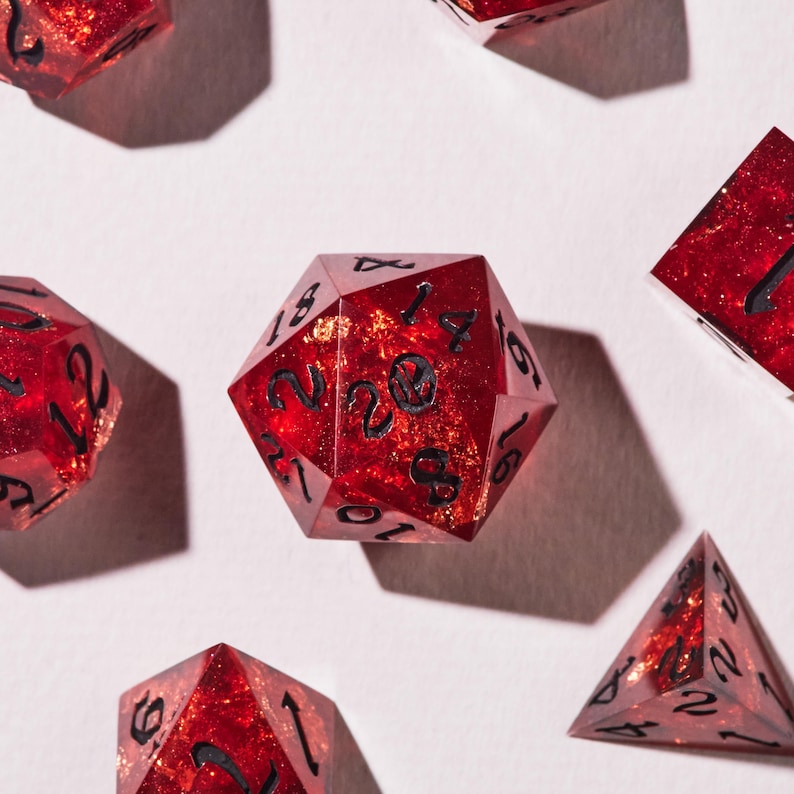 May include: A selection of red, multi-sided dice with black numbering. The dice have a glittery, speckled appearance. The dice are arranged on a white surface, with shadows cast from an overhead light source. The dice are used for tabletop games.