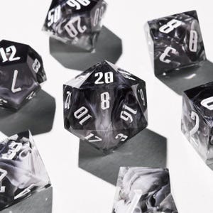 Puede incluir: Un conjunto de dados poliédricos en blanco y negro, incluyendo un dado de veinte caras. Los dados tienen números blancos y un efecto veteado. Los dados están colocados sobre una superficie blanca.