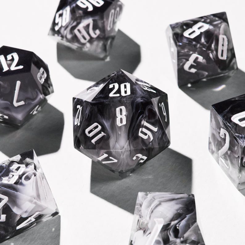 May include: A selection of black and white marbled polyhedral dice, including a twenty-sided die. The dice have white numbering. The dice are displayed on a white surface, with shadows cast.
