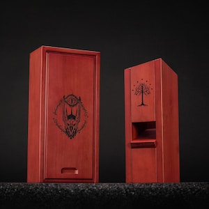 May include: Two wooden boxes with a reddish-brown finish. One box is rectangular with a drawer and a black graphic of a menacing eye and inscription. The other box is a tall, open-front design with a tree graphic.