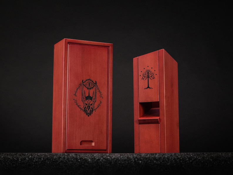 May include: Two wooden boxes with a reddish-brown finish. One box is rectangular with a drawer and a black graphic of a menacing eye and inscription. The other box is a tall, open-front design with a tree graphic.