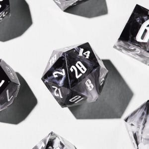 May include: Several polyhedral dice, including a black and white d20 with white numerals. The dice have a marbled effect and are arranged on a white surface, with shadows cast beneath them.