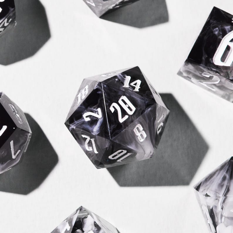 May include: Several polyhedral dice, including a black and white d20 with white numerals. The dice have a marbled effect and are arranged on a white surface, with shadows cast beneath them.