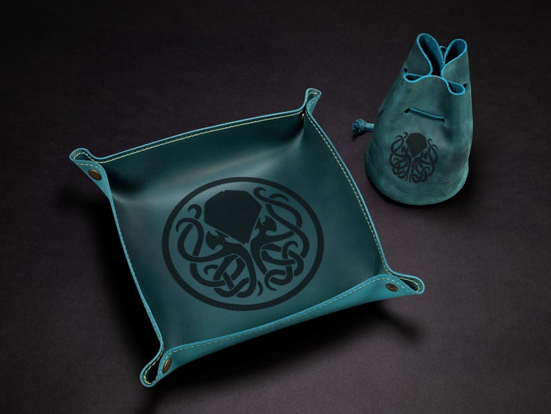 May include: Teal leather tray and drawstring pouch set. The tray has a square shape with rounded corners and a black octopus design. The pouch has a matching octopus design. The set is made of leather.