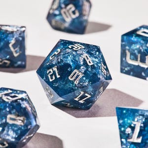 Könnte beinhalten: Eine Sammlung von polyedrischen Würfeln mit blauem und silbernem Marmordesign. Die Würfel haben verschiedene Formen, darunter ein 20-seitiger Würfel, und sind mit silbernen Zahlen versehen. Die Würfel sind vor einem weißen Hintergrund angeordnet.