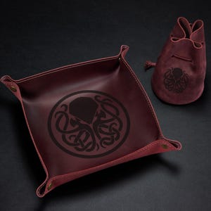May include: Burgundy leather dice tray and drawstring bag set. The tray is square with snap closures at the corners and a black octopus design. The bag also features the octopus design.