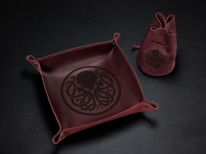 May include: Burgundy leather dice tray and drawstring bag set. The tray is square with snap closures at the corners and a black octopus design. The bag also features the octopus design.