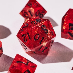 May include: A collection of red, multi-sided dice with black numerals and gold flecks. The dice are transparent, with the gold flecks visible. These dice are used for tabletop games and are set against a white background.