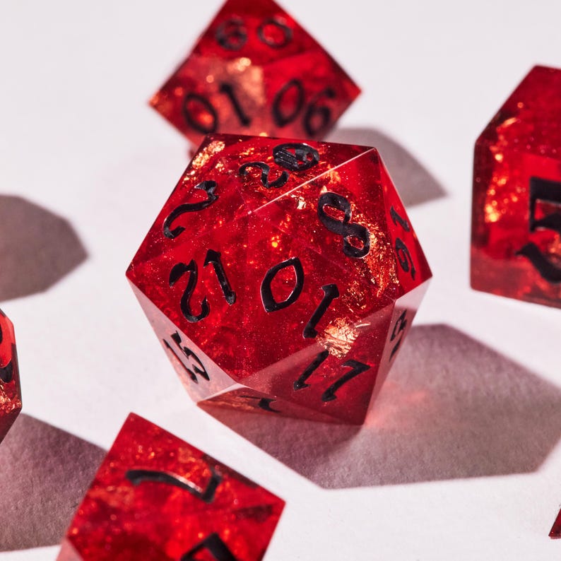 May include: A collection of red, multi-sided dice with black numerals and gold flecks. The dice are transparent, with the gold flecks visible. These dice are used for tabletop games and are set against a white background.