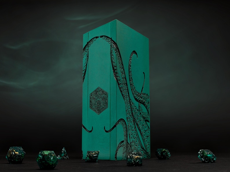 May include: A teal dice box with black tentacles and geometric designs. Several green and gold dice are scattered in front of the box. The box is rectangular and stands upright against a dark background.