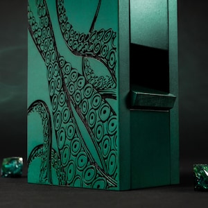 May include: A dark teal dice box with a black octopus tentacle design. The box has a rectangular shape with a small, rectangular opening. Several dice with green and teal accents are scattered around the box.