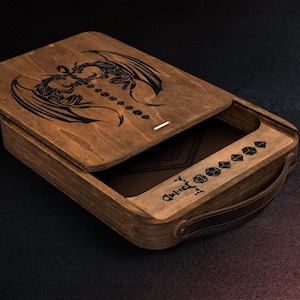 Puede incluir: Una caja de madera con tapa con bisagras, con un diseño de dragón grabado con láser. La caja está abierta, revelando un compartimento con símbolos de dados y un asa de cuero. La madera tiene un tono marrón cálido.