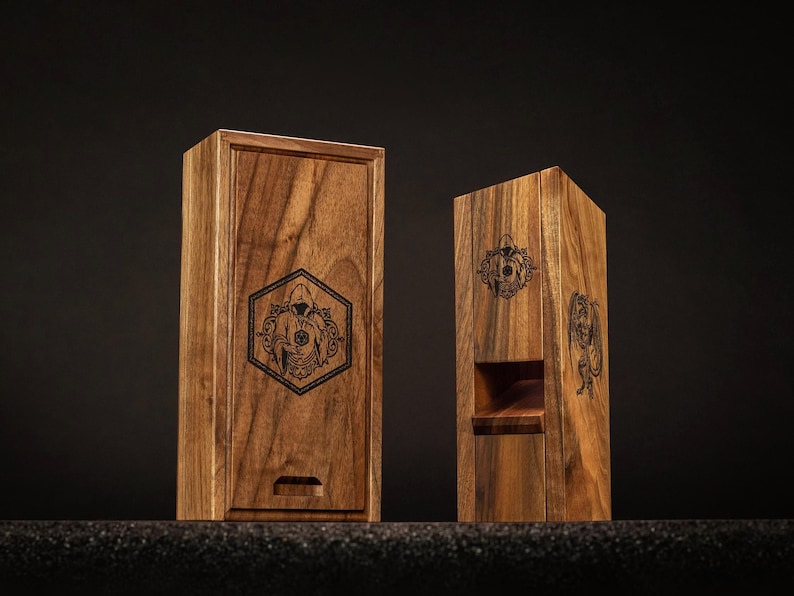 May include: Two wooden boxes with engraved designs. One is rectangular with a sliding lid, the other is a tall, open-front box. Both are made of wood with a natural grain and feature a dark, detailed graphic.