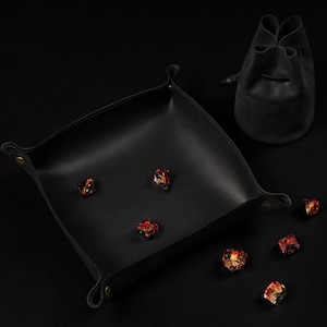 May include: A black leather dice tray with rounded corners and a matching drawstring bag. Several multi-colored dice are scattered on the tray and surface, creating a gaming theme.