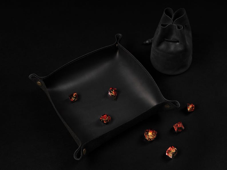 May include: A black leather dice tray with rounded corners and a matching drawstring bag. Several multi-colored dice are scattered on the tray and surface, creating a gaming theme.