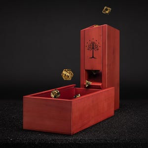 May include: A red wooden dice tower and tray set with a tree emblem. Several gold-colored dice are scattered around the tower and tray. The tower has a rectangular shape with a slot for dice to roll out.