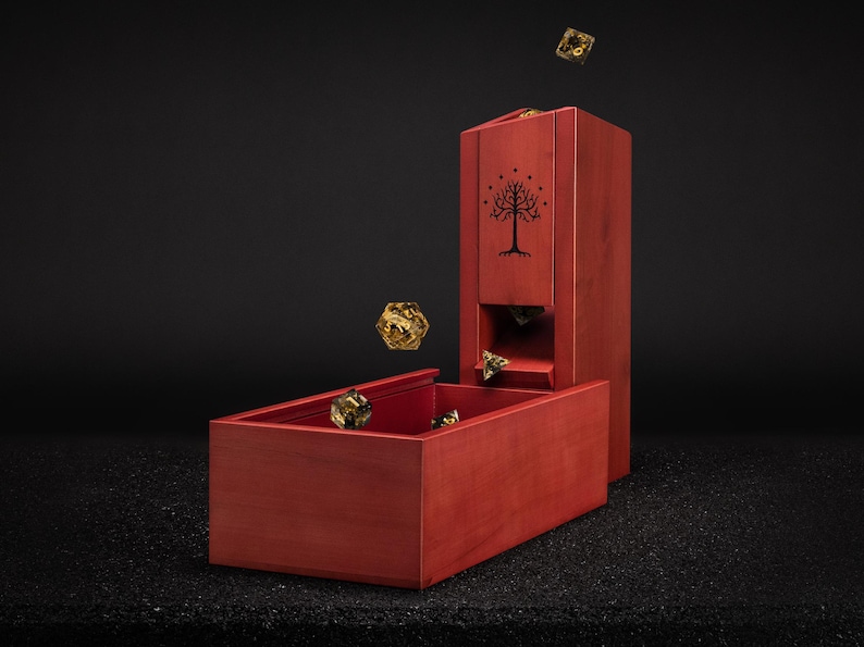 May include: A red wooden dice tower and tray set with a tree emblem. Several gold-colored dice are scattered around the tower and tray. The tower has a rectangular shape with a slot for dice to roll out.