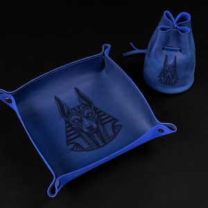May include: A blue leather tray and drawstring pouch set. Both items feature a dark embossed image of Anubis, the Egyptian god of the afterlife. The tray has a square shape with rounded corners and snap closures. The pouch is cylindrical.