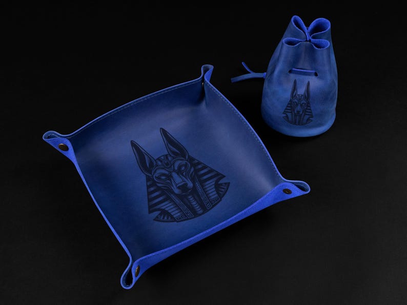 May include: A blue leather tray and drawstring pouch set. Both items feature a dark embossed image of Anubis, the Egyptian god of the afterlife. The tray has a square shape with rounded corners and snap closures. The pouch is cylindrical.