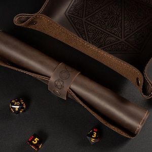 May include: A brown leather dice tray and a rolled-up dice holder with a strap featuring engraved dice symbols. Several multi-sided dice in red, black, and gold are scattered around the tray and holder. The tray has a large engraved 20.