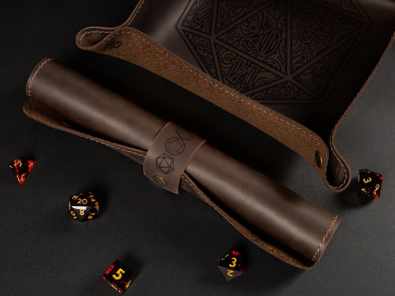 May include: A brown leather dice tray and a rolled-up dice holder with a strap featuring engraved dice symbols. Several multi-sided dice in red, black, and gold are scattered around the tray and holder. The tray has a large engraved 20.