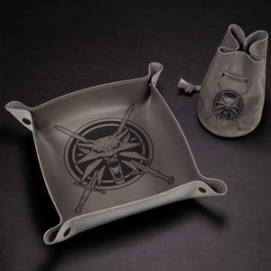 May include: Gray leather tray and pouch set. The tray is square with rounded corners and a raised edge. Both items feature a black graphic of a wolf's head with crossed swords. The pouch has a drawstring closure.