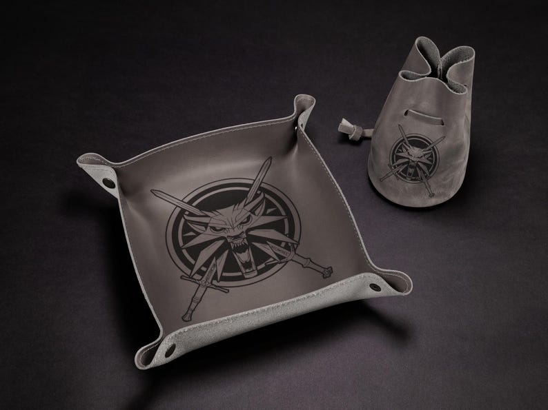 May include: Gray leather tray and pouch set. The tray is square with rounded corners and a raised edge. Both items feature a black graphic of a wolf's head with crossed swords. The pouch has a drawstring closure.