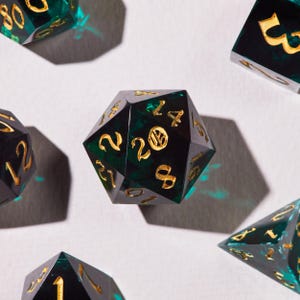 May include: Close-up of several polyhedral dice. The dice are black with a green, swirling interior and gold numbering. The dice are arranged on a white surface, with shadows cast by the lighting.