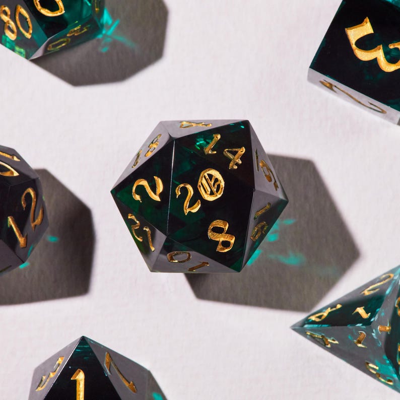 May include: Close-up of several polyhedral dice. The dice are black with a green, swirling interior and gold numbering. The dice are arranged on a white surface, with shadows cast by the lighting.