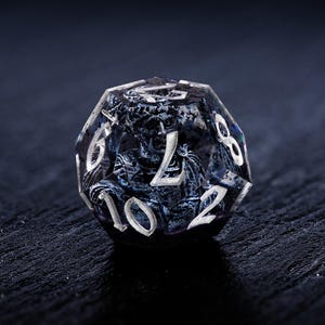 K&ouml;nnte beinhalten: Ein klarer, vielseitiger W&uuml;rfel mit detailliertem, dunkelblauem und grauem Innendesign. Der W&uuml;rfel zeigt eine geformte Figur und wei&szlig;e Zahlen. Ideal f&uuml;r Tabletop-Spiele, dieser einzigartige W&uuml;rfel verleiht jedem Spiel einen Hauch von Fantasie.