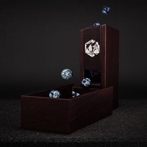 May include: A dark wood dice tower and tray set with a geometric design. Several blue and white dice are shown in the tray, falling from the tower, and in mid-air. The tower has a decorative emblem on the front.