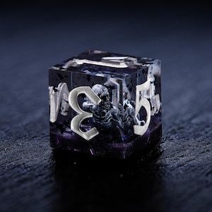 May include: A clear, cube-shaped dice with a silver number "3" and a silver number "5" on the visible sides. The dice is filled with a black and white resin with a skeletal figure inside.
