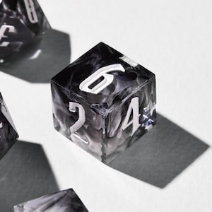 May include: A close-up of a six-sided die with white numbers on a black and grey marbled background. The die is casting a shadow on a white surface. Other dice are visible in the background.