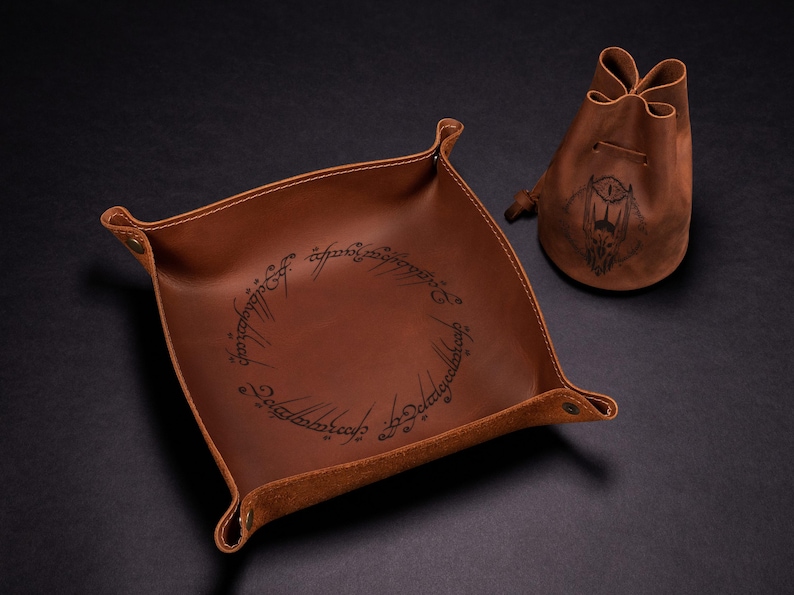 May include: A brown leather tray and drawstring pouch set. The square tray has rounded corners and a stitched border. The tray and pouch feature a design from a fantasy novel. The pouch has a graphic of an eye.