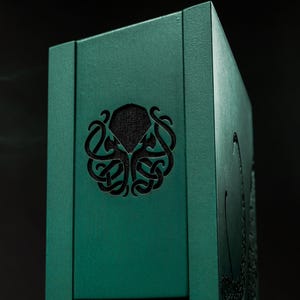 May include: A teal wooden box featuring a carved octopus design. The box has a dark, textured interior visible through the cut-out design. The box is rectangular and set against a dark background.