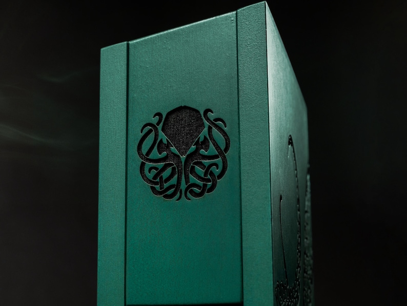 May include: A teal wooden box featuring a carved octopus design. The box has a dark, textured interior visible through the cut-out design. The box is rectangular and set against a dark background.