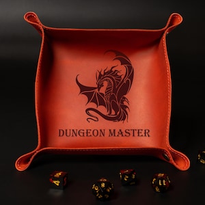 May include: An orange leather dice tray with a dragon illustration and the text "DUNGEON MASTER". The tray has a square shape with curved edges and is surrounded by several dice.