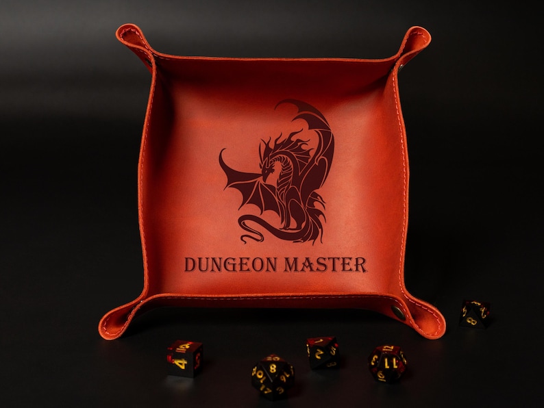 May include: An orange leather dice tray with a dragon illustration and the text "DUNGEON MASTER". The tray has a square shape with curved edges and is surrounded by several dice.