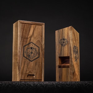 May include: Two wooden boxes with engraved designs. The box on the left is rectangular with a sliding lid and a hexagonal emblem. The box on the right is a tall, open-front design with a similar emblem. Both are made of dark wood.