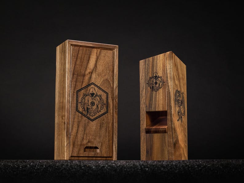 May include: Two wooden boxes with engraved designs. The box on the left is rectangular with a sliding lid and a hexagonal emblem. The box on the right is a tall, open-front design with a similar emblem. Both are made of dark wood.