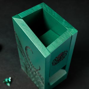 May include: A teal wooden dice box with engraved tentacles and a symbol. The box is rectangular with a recessed top and a small opening on one side. Three teal dice are scattered around the box on a black surface.