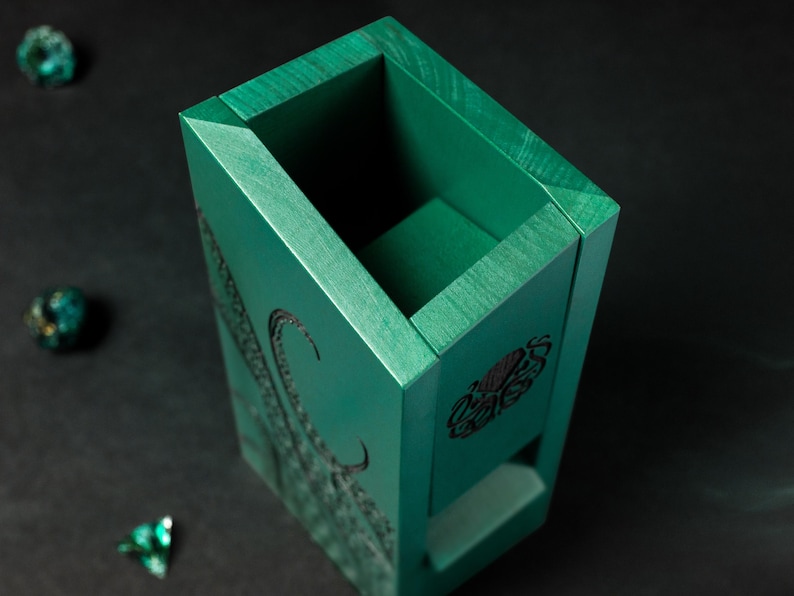 May include: A teal wooden dice box with engraved tentacles and a symbol. The box is rectangular with a recessed top and a small opening on one side. Three teal dice are scattered around the box on a black surface.
