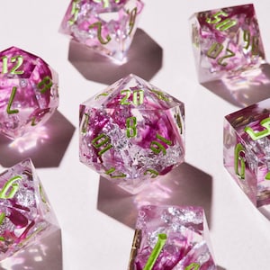 May include: A collection of clear, multi-sided dice with a floral design. The dice are filled with pink and silver accents, and the numbers are in a bright green color. The dice are scattered on a white surface.