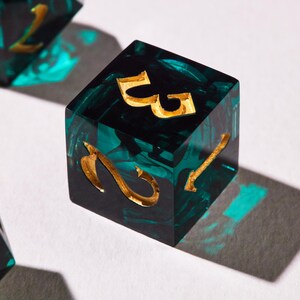 May include: A close-up of a six-sided die with gold-coloured numbers. The die is a mix of black and teal green, with a marbled effect. The numbers are etched into the die. The die is sitting on a white surface.