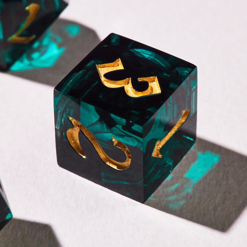 May include: A close-up of a six-sided die with gold-coloured numbers. The die is a mix of black and teal green, with a marbled effect. The numbers are etched into the die. The die is sitting on a white surface.