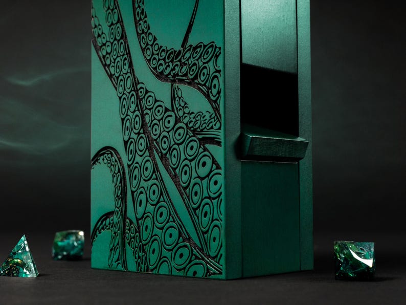 May include: A dark teal dice box featuring a black octopus tentacle design. The box is rectangular with a small opening. Several dice with green and teal accents are scattered around the box.