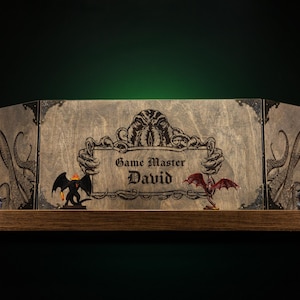 Może przedstawiać: Drewniany ekran mistrza gry z grawerowanymi grafikami i napisem "Game Master David". Ekran przedstawia ilustracje macek, smok&oacute;w i stworzenia. Ekran jest eksponowany na drewnianej p&oacute;łce.