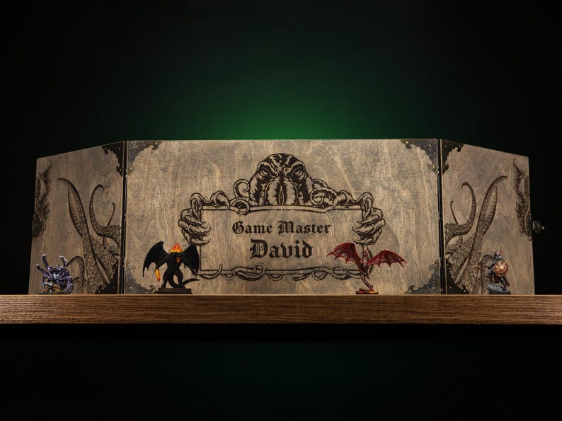 Może przedstawiać: Drewniany ekran mistrza gry z grawerowanymi grafikami i napisem "Game Master David". Ekran przedstawia ilustracje macek, smok&oacute;w i stworzenia. Ekran jest eksponowany na drewnianej p&oacute;łce.