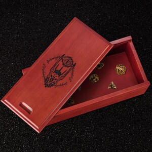 May include: A red wooden dice box with a hinged lid, featuring a black graphic design and text. The box is open, revealing several dice in various shapes and colors. The dice box is on a black surface.
